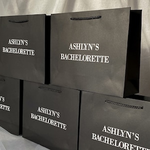 May include: Stack of black paper gift bags with white text that reads "Ashlyn's Bachelorette".