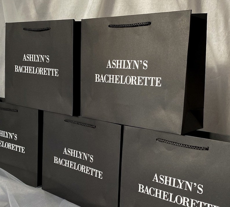 May include: Stack of black paper gift bags with white text that reads "Ashlyn's Bachelorette".