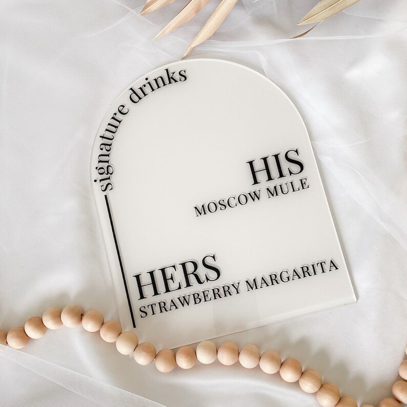 May include: White acrylic sign with black text that reads "signature drinks". The sign is shaped like an arch and has two drink options: "HIS Moscow Mule" and "HERS Strawberry Margarita".