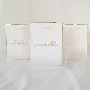 May include: Three white gift bags with gold trim and white handles. The bags are labeled with the names "Christina", "Samantha", and "Nicole" in gold lettering.
