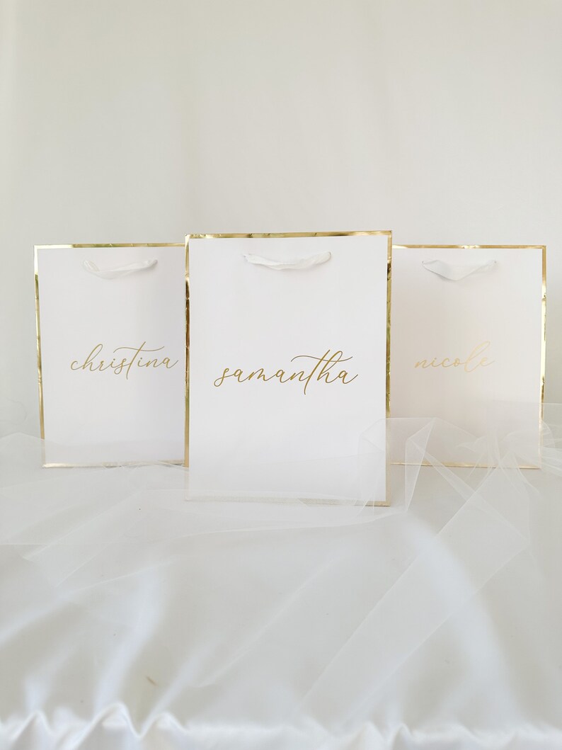 May include: Three white gift bags with gold trim and white handles. The bags are labeled with the names "Christina", "Samantha", and "Nicole" in gold lettering.