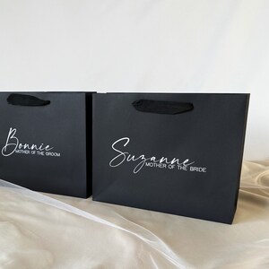 May include: Two black gift bags with black ribbon handles. The bag on the left has the name "Bonnie" and the text "Mother of the Groom" printed in white. The bag on the right has the name "Suzanne" and the text "Mother of the Bride" printed in white.