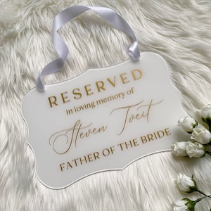 May include: White acrylic sign with gold lettering that reads "Reserved in loving memory of Steven Tveit Father of the Bride". The sign has a white ribbon attached to the top for hanging.