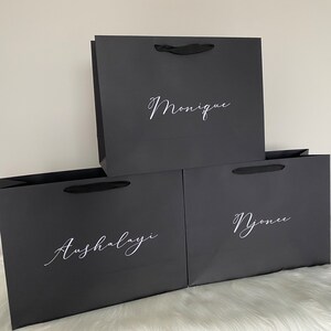 May include: Three black gift bags with white cursive text reading "Monique", "Aushalayi", and "Njonee". Each bag features a black ribbon handle. The bags are displayed on a white surface.