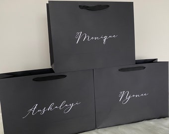 以下が含まれることがあります： 「Monique」、「Aushalayi」、「Njonee」と書かれた白い筆記体の文字が特徴の、黒いギフトバッグが3つ。各バッグには黒いリボンハンドルが付いています。白い表面に配置されています。