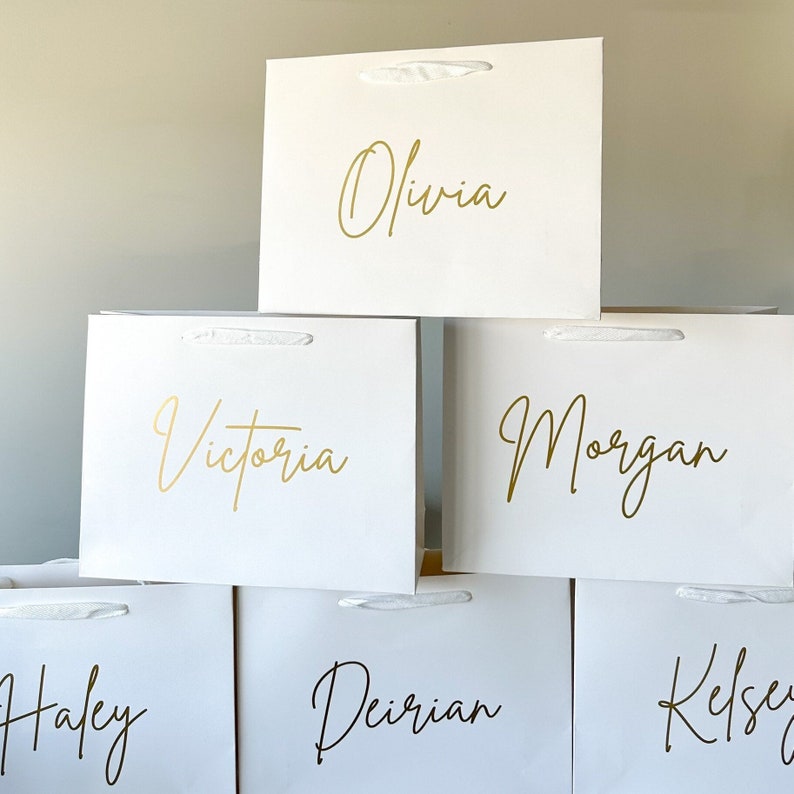 May include: Six white gift bags with gold lettering. The names Olivia, Victoria, Morgan, Haley, Deiriian, and Kelsey are written on the bags.