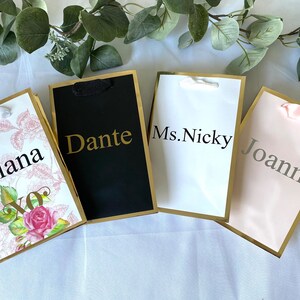 May include: Four gift bags with gold trim and different designs. The bags are labeled with the names "Giana", "Dante", "Ms. Nicky", and "Joann".