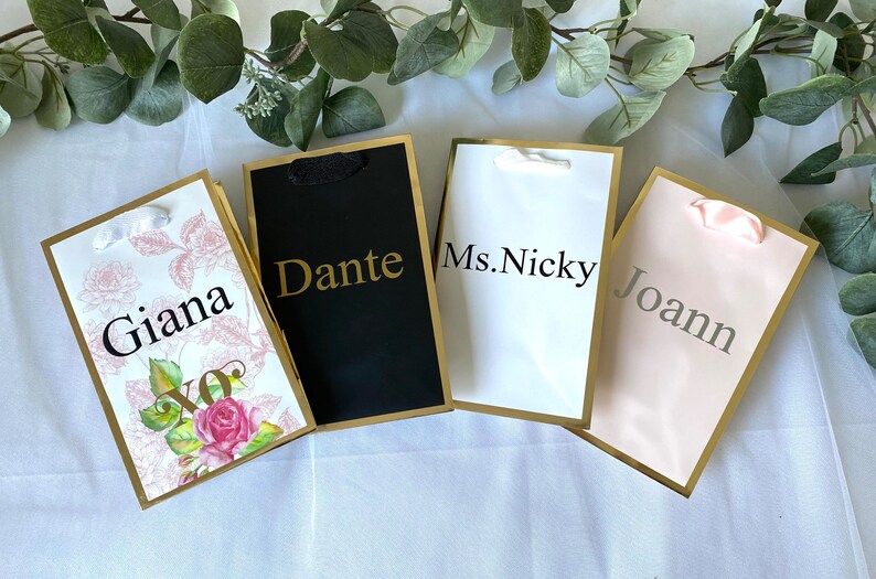 May include: Four gift bags with gold trim and different designs. The bags are labeled with the names "Giana", "Dante", "Ms. Nicky", and "Joann".
