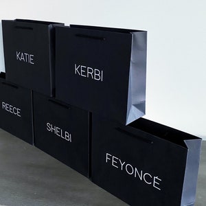 May include: Five black paper shopping bags stacked on top of each other. The bags have white text on them. The text on the bags reads "KATIE", "KERBI", "REECE", "SHELBI", and "FEYONC&Eacute;".