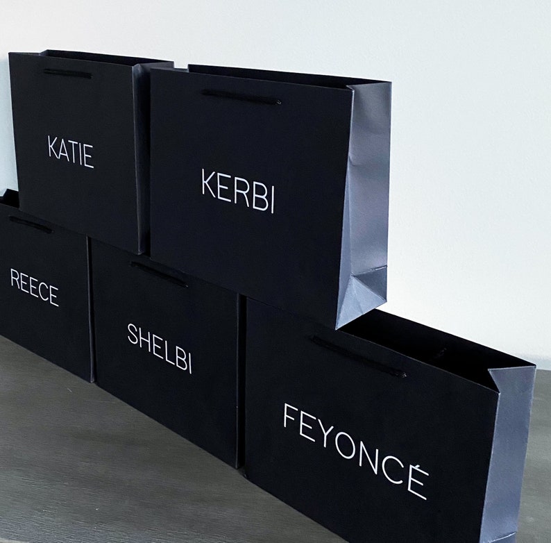 May include: Five black paper shopping bags stacked on top of each other. The bags have white text on them. The text on the bags reads "KATIE", "KERBI", "REECE", "SHELBI", and "FEYONC&Eacute;".