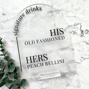 May include: Clear acrylic sign with rounded top and black text that reads "signature drinks". The sign lists two drinks: "HIS OLD FASHIONED" and "HERS PEACH BELLINI".