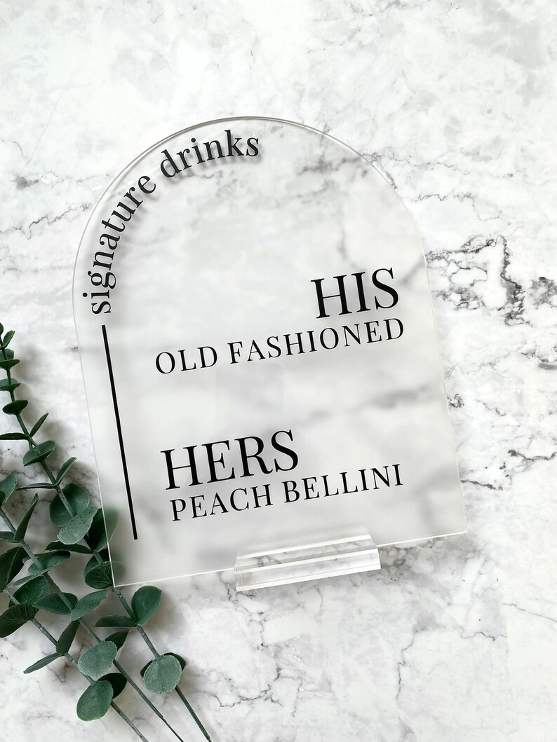 May include: Clear acrylic sign with rounded top and black text that reads "signature drinks". The sign lists two drinks: "HIS OLD FASHIONED" and "HERS PEACH BELLINI".