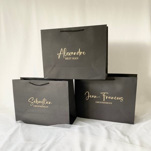 May include: Three black gift bags with gold lettering. The top bag says "Alexandre Best Man". The middle bag says "S&eacute;bastien Groomsman". The bottom bag says "Jean-Francois Groomsman".