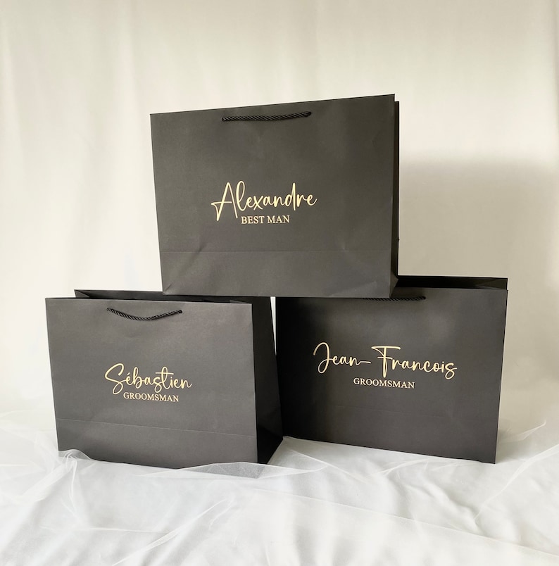 May include: Three black gift bags with gold lettering. The top bag says "Alexandre Best Man". The middle bag says "S&eacute;bastien Groomsman". The bottom bag says "Jean-Francois Groomsman".
