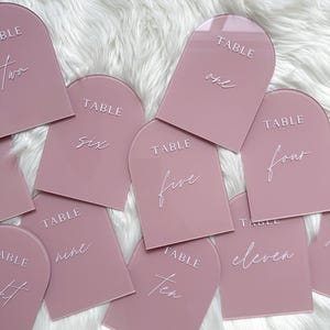 May include: A set of ten pink acrylic table numbers with a rounded arch top. The numbers are written in white script font and the word "TABLE" is printed above each number.
