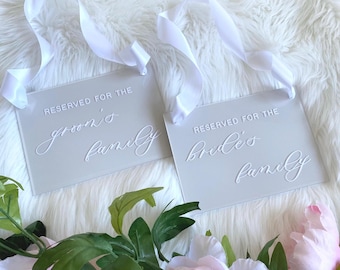 May include: Two gray acrylic signs with white ribbon loops. The signs read "Reserved for the groom's family" and "Reserved for the bride's family".