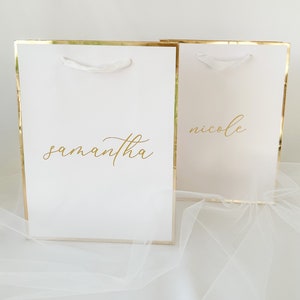 May include: Two white gift bags with gold trim and white handles. The bags have the names "Samantha" and "Nicole" written in gold lettering.