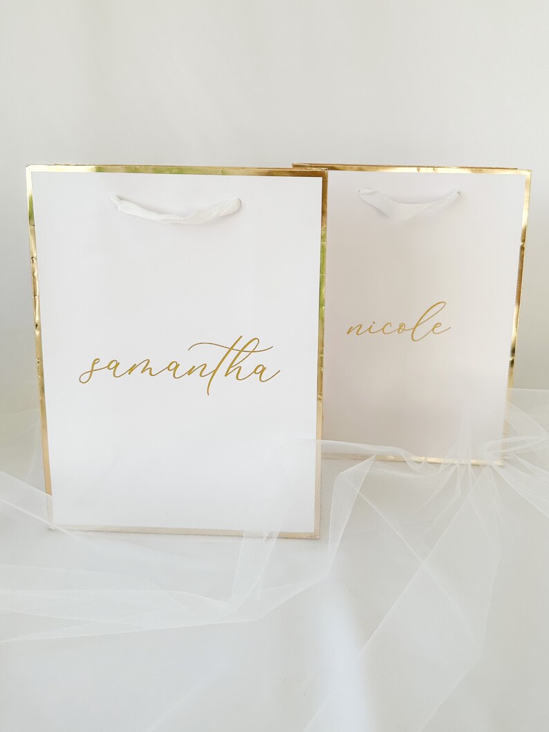 May include: Two white gift bags with gold trim and white handles. The bags have the names "Samantha" and "Nicole" written in gold lettering.