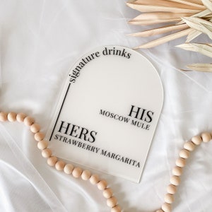 May include: White acrylic sign with rounded top, featuring the text "signature drinks" in black. The sign is divided into two sections, one labeled "HIS" with the text "MOSCOW MULE" and the other labeled "HERS" with the text "STRAWBERRY MARGARITA".