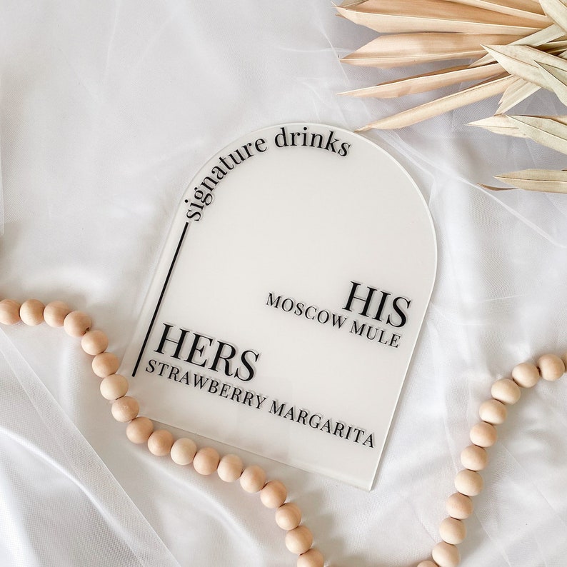 May include: White acrylic sign with rounded top, featuring the text "signature drinks" in black. The sign is divided into two sections, one labeled "HIS" with the text "MOSCOW MULE" and the other labeled "HERS" with the text "STRAWBERRY MARGARITA".