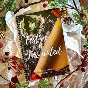 May include: A black spiral bound cookbook with the title "Festive & Fermented" and a photo of a rolling pin on a floured surface with a green cookie cutter in the shape of a Christmas tree. The cookbook is by Laura Bruner, the Sourdough Sisterhood.