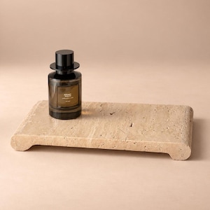 May include: A cylindrical glass perfume bottle with a black cap and a label reading EBONY WOOD ZARA EMOTIONS ELIXIR, positioned on a rectangular, beige, porous stone tray with rounded edges and a raised platform design.
