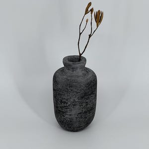 May include: A small, textured, dark grey vase with a rounded body and a narrow neck. A dried, brown twig with small seed pods is placed inside the vase. The vase is set against a plain white background.