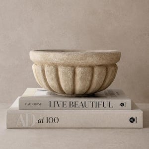 May include: A beige, fluted, stone-like bowl with a rounded rim sits atop two stacked books. The top book is titled LIVE BEAUTIFUL by Calderone, and the bottom book is titled AD at 100. The bowl has a textured, matte finish and vertical grooves.