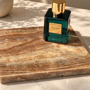 Op de afbeelding: Een vierkante parfumfles van diep smaragdgroen glas met een glanzende, cilindervormige gouden dop. De fles heeft een rechthoekig goudkleurig etiket aan de voorzijde met de tekst "FANCIER" en "EAU DE PARFUM" in het zwart gedrukt, rustend op een bruine stenen plaat.