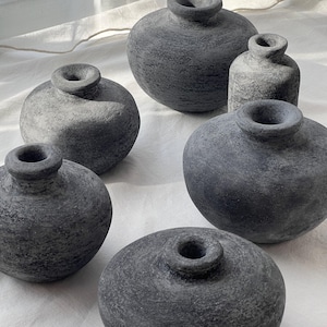 Puede incluir: Una colección de cinco jarrones pequeños de cerámica gris oscuro con una textura rugosa similar a la piedra. Cada jarrón presenta un cuerpo redondeado y una abertura circular estrecha en la parte superior. Los recipientes varían en tamaño.