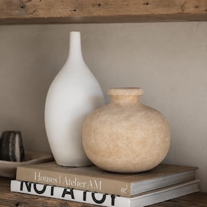 Op de afbeelding: Een hoge, gladde witte keramische vaas met een smalle hals staat naast een kortere, ronde, getextureerde beige vaas met een brede opening. Beide vazen staan op twee gestapelde boeken met de titels Houses | Atelier AM en NOT A TOY.