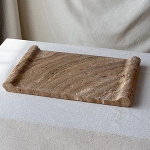 May include: A rectangular brown stone tray featuring a natural porous texture and visible mineral veining. The tray has two raised, rounded edges on opposite sides, creating a concave surface with a distinct earthy appearance and organic patterns.