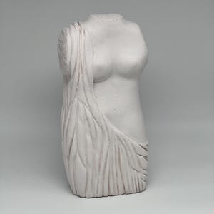 May include: A white plaster sculpture of a female torso. The upper body is depicted from the chest up, with draped fabric over the left shoulder and side. The sculpture has a classical design and aesthetic.