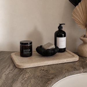 Op de afbeelding: Een rechthoekig dienblad van beige travertijn met een zwarte pot Palermo bodyscrub, een grijs stuk zeep in een zwart bubbelvormig schaaltje en een zwarte pompflacon met het label L'ASCARI handzeep, geplaatst op een stenen aanrecht.