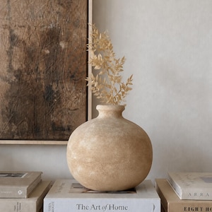 Puede incluir: Un jarrón de cerámica beige, redondo y texturizado, con un cuello estrecho y un cuerpo esférico ancho. El jarrón contiene una rama seca de color tostado claro. Está colocado sobre un libro blanco titulado The Art of Home de Shea McGee.
