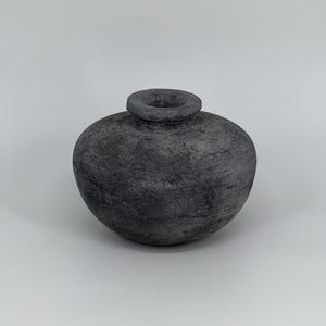 May include: A dark grey, round ceramic vase with a textured surface. The vase has a small, circular opening at the top and a bulbous body. The vase is set against a plain, light background.