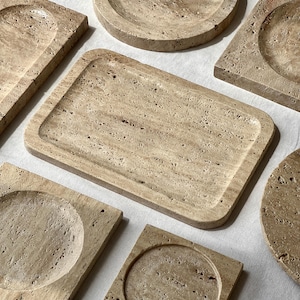 May include: A collection of beige travertine stone trays and coasters in various shapes, including rectangular, square, and circular designs. Each piece features a porous, textured surface with natural variations in color and stone grain patterns.