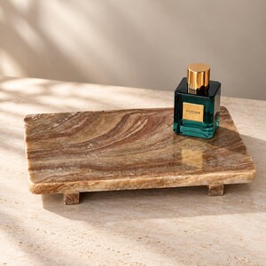 Puede incluir: Una bandeja rectangular de piedra veteada en tonos marrones y tostados con patas cortas y superficie pulida. Un frasco de perfume cuadrado de cristal color verde azulado con tapón dorado y la etiqueta FANCIER se encuentra sobre la bandeja.