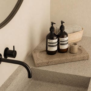 May include: Two amber glass pump bottles labeled Aesop sit on a rectangular, textured stone tray alongside a small cylindrical candle. The bottles feature black pump dispensers and white labels with black text, positioned near a matte black faucet.