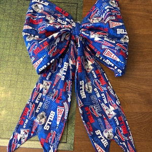 May include: A large blue fabric bow with a repeating pattern of the word "BILLS" in white and red, and the team's logo. The bow is likely for decoration or as a hair accessory. The bow is on a wooden surface. The bow's dimensions are not visible.