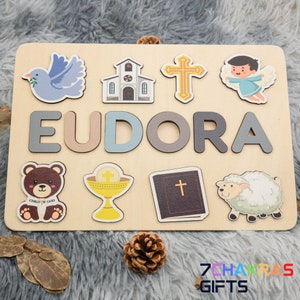 May include: A wooden personalized puzzle with the name Eudora and religious imagery, including a dove, church, cross, angel, bear, chalice, bible, and lamb. The puzzle is on a light brown wooden background.