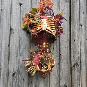 May include: A gold-colored skeleton ribcage decorated with fall-colored flowers and leaves. The flowers are in shades of red, orange, purple, and brown. The skeleton is attached to a wooden surface.