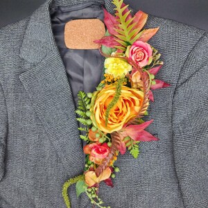 May include: A gray plaid blazer adorned with a vibrant floral arrangement. The arrangement features orange and pink roses, yellow flowers, and various green and red leaves. A cork piece is pinned to the blazer's lapel.