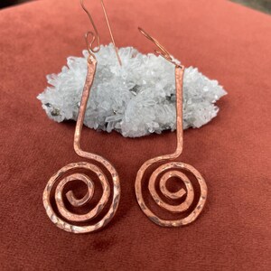 May include: A pair of copper earrings featuring a hammered texture and a spiral design. Each earring has a long, straight stem leading into a circular coil at the bottom, finished with a simple hook wire for attachment to the earlobe.