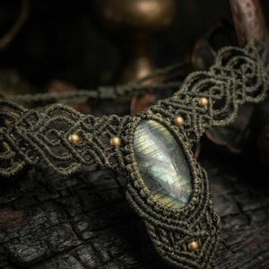 Può includere: Collana in macramè fatta a mano con una grande pietra di labradorite ovale. La collana è realizzata con cordoncino verde oliva e impreziosita da piccole perline dorate. L'intricato nodo crea un design dettagliato e strutturato, perfetto per stili bohémien.