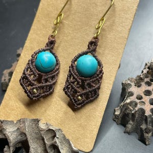May include: A pair of dangle earrings featuring round turquoise beads set within dark brown macrame cord patterns. Each earring hangs from a gold-toned metal hook and includes a small gold-toned bead accent at the bottom of the woven design.