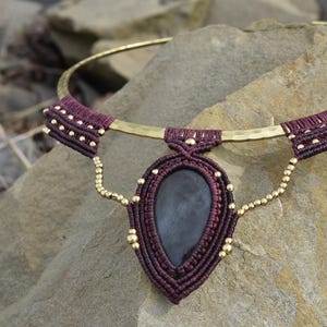 Op de afbeelding: Een handgemaakte ketting met een donkerrood macrame-ontwerp met goudkleurige accenten. De ketting heeft een druppelvormige hanger met een donkere stenen kern en een gehamerde goudkleurige metalen kraag. De ketting wordt op een rotsachtig oppervlak getoond.