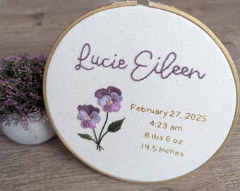 May include: Embroidered hoop with the name "Lucie Eileen" in cursive script, a floral design with purple and pink flowers, and the text "February 27, 2025 4:23 am 8 lbs 6 oz 19.5 inches".