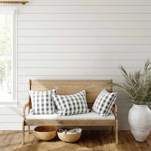 Puede incluir: Un banco de madera con un cojín a cuadros blanco y gris y tres almohadas. El banco está frente a una pared blanca de tablones de madera y un jarrón con vegetación seca.