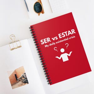 以下が含まれることがあります： 「SER vs ESTAR My daily existential crisis」の白文字が入った赤いリングノート。ノートには、疑問符が付いた人物のシンプルな白いグラフィックが描かれています。白い表面には、サングラスやゴールドのバインダークリップなどのアイテムがあります。